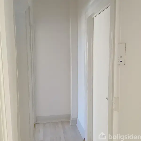 En hvidmalet gang med to døråbninger fører til et tomt rum. Lyset skaber en ren, minimalistisk atmosfære. Ingen tekst fundet i billedet.