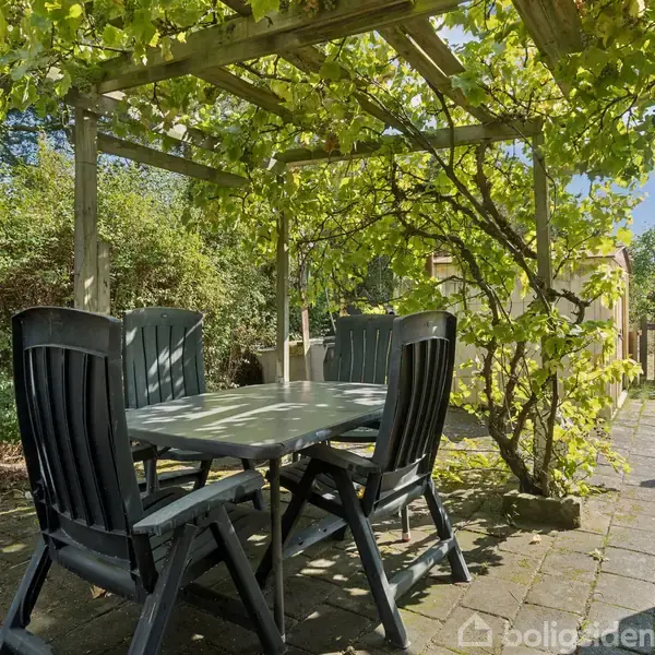 Et havebord omgivet af fire sorte stole står under en pergola med grønne planter. Omkring er der en stenbelagt terrasse og grønne buske.