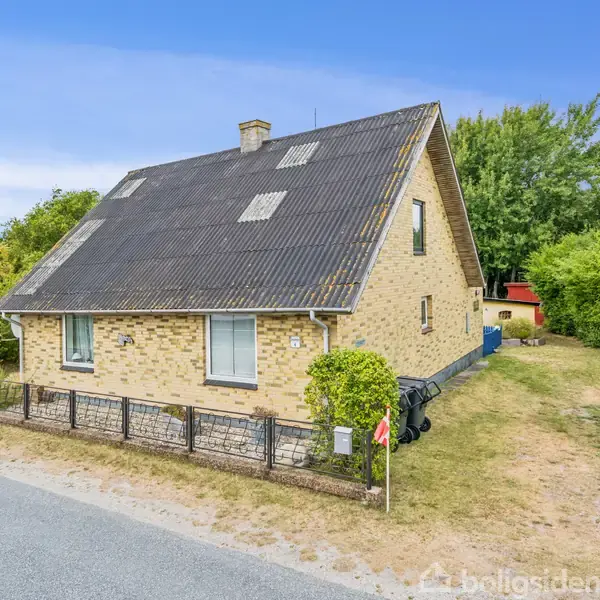 Et gult murstenshus med skråt tag står ved vejen på en ujævn grund. Omkring huset er der buske og træer, og en lav hæk omkranser det forreste området.