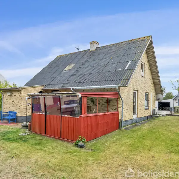 Et murstenshus med sort tag har en rød udestue. Det står i en have med en græsplæne, træer og en blå bænk ved siden af terrassen.