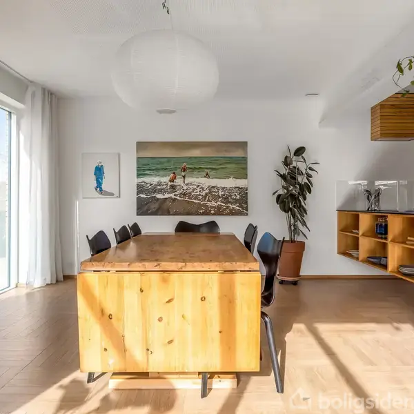 Et langt træbord omgivet af sorte stole står i et lyst spisestueområde med en stor vinduesfacade. Væggene er dekoreret med kunst, og planter pryder rummet.