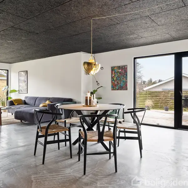 Stue med rund spisebord og fire stole, står i lys stue med gråt hjørnesofa. Skydedør åbner til terrasse og have. Moderne indretning med planter og kunstværker på væggene.