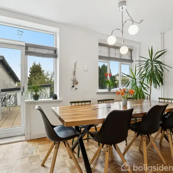 Et spisebord med seks sorte stole står i et lyst rum med trægulv. En plante og blomster dekorerer bordet ved et vindue med udsigt til en terrasse og træer udenfor.