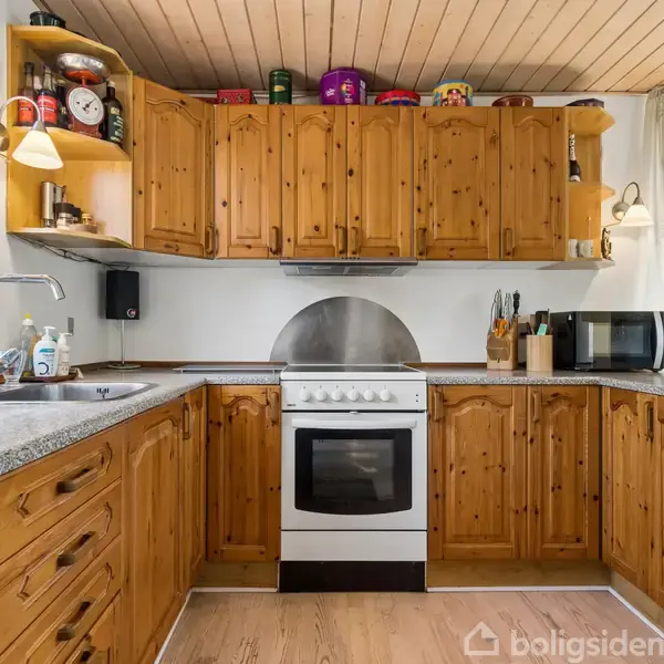 Køkken med træskabe omfavner en hvid komfur. Køkkenredskaber står på bordpladen. Et stort vindue med planter til venstre og et hvidt køleskab til højre tilfører lys.
