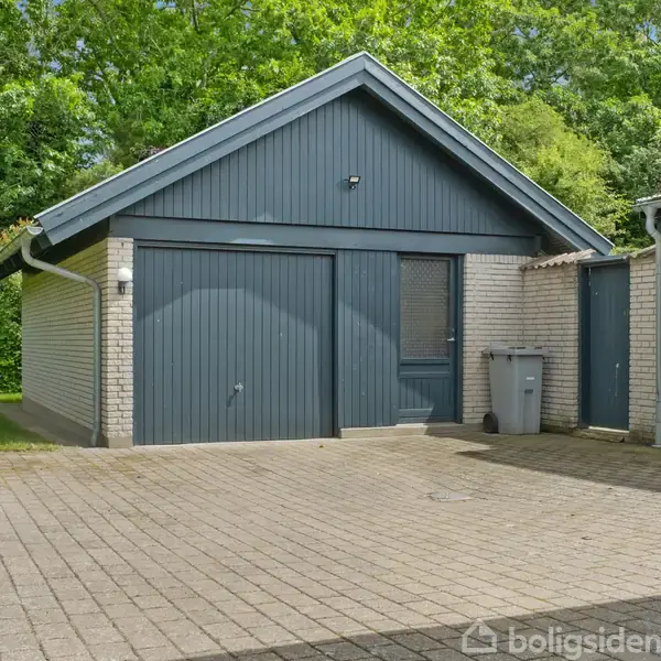 Garage med blå dør og tilstødende skur. Aktionsløs og omgivet af grønne buske og træer. Foran er der en flisebelagt indkørsel og en skraldespand står ved siden af bygningen.