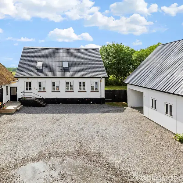 Et hvidt, traditionelt dansk hus med gråt tag står stille i midten af en grusbelagt gårdsplads, omgivet af to mindre bygninger. Bagved ses grønne træer og en blå himmel.