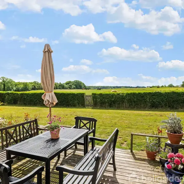 En sort havemøbelgruppe står stille på en terrasse med udsigt til en grøn græsplæne og en hæk. Der er blomsterkrukker og en lukket parasol på bordet.
