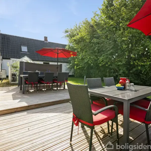 Terrassemøbler står på en træterrasse med røde parasoller og stolehynder, omgivet af en have med græs og buske. Et hus med gråt tag og hvide vægge ses i baggrunden.