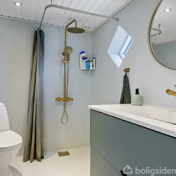 Brusebad med guldbruser og hvid toilet, placeret i et moderne badeværelse med lyse vægge og rund spejl. Ved siden af vasken står en dispenser og et håndklæde.