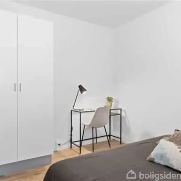Et værelse har en seng med brun dyne, et lille skrivebord med lampe og plante, og et hvidt garderobeskab. Gulvet er af træplanker.