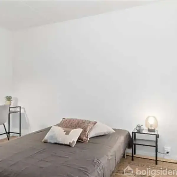 Soveværelse med enkeltseng dækket af puder, stående centralt. Et skrivebord med lampe og stol står i hjørnet. En sidebordslampe lyser ved siden af sengen. Ingen tekst.