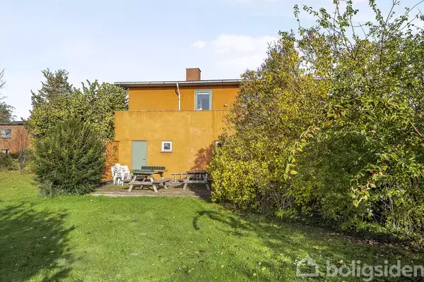 Et orange hus med fladt tag står omgivet af grønne buske og græs. Foran huset er der et picnicbord og nogle hvide plaststole.