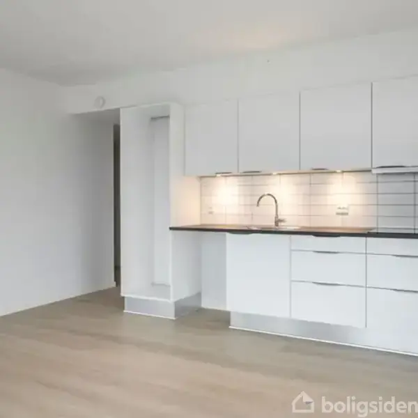 Et stilrent køkken med hvide skabe og skuffer, en indbygget ovn og en moderne vandhane over en sort bordplade. Rummet har lyse trægulve og en minimalistisk indretning.
