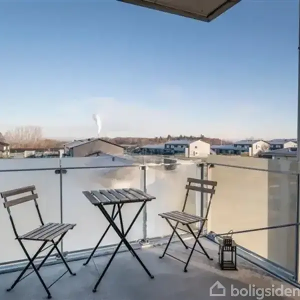 To klapstole og et lille bord står på en balkon med glasrækværk. I baggrunden ses et boligområde under en blå himmel. En lanterne står ved siden af stolene.