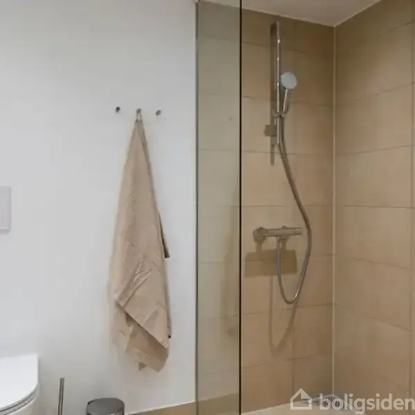 Et badeværelse har en vægmonteret bruser med glasafskærmning i et rum med lyse fliser. Ved siden af hænger et beige håndklæde på en krog. Toilettet er hvidt.