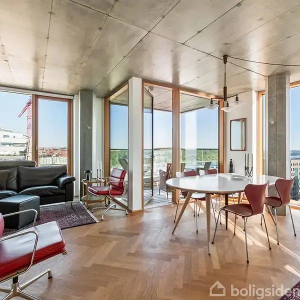 En moderne stue med røde stole omkring et rundt, hvidt bord. Store vinduer giver udsigt til byen og havet. Trægulvet kombinerer med gråt loft i et stilrent design.