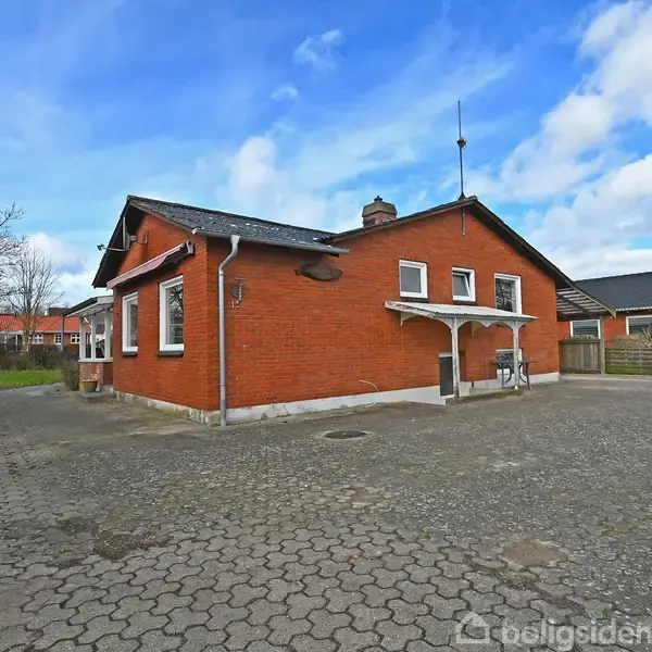 Rødt murstenshus med hvide vinduesrammer står på en flisebelagt gårdsplads. Omkring bygningen er der enkelte træer og buske, under en blå himmel med skyer.