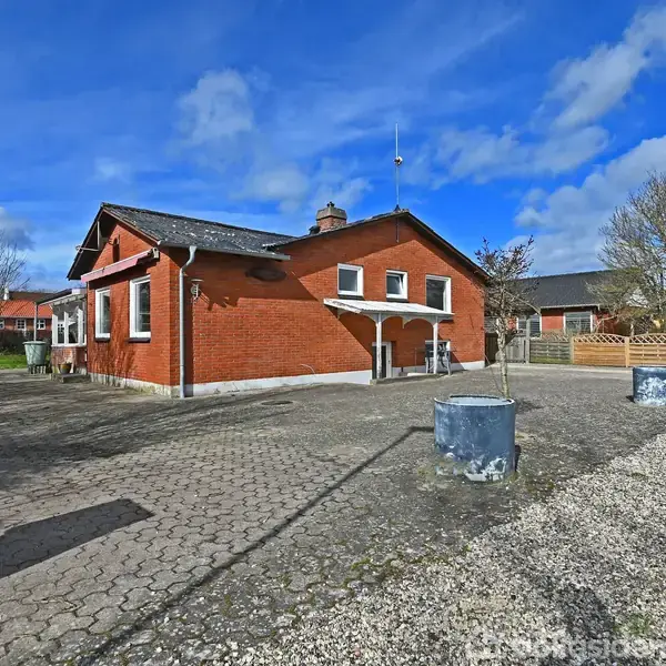 Rødt murstenshus med sort tag står stille på flisebelagt grund. Sollys skinner på facaden. Omgivet af træer og hegn i et roligt boligkvarter under en blå himmel med skyer.