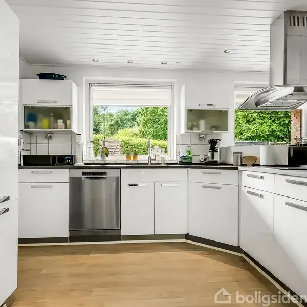 Rækkehus med rødt tegltag står stille bag en grøn hæk. Foran huset er en terrasse med sorte møbler. I baggrunden ses frodige træer.