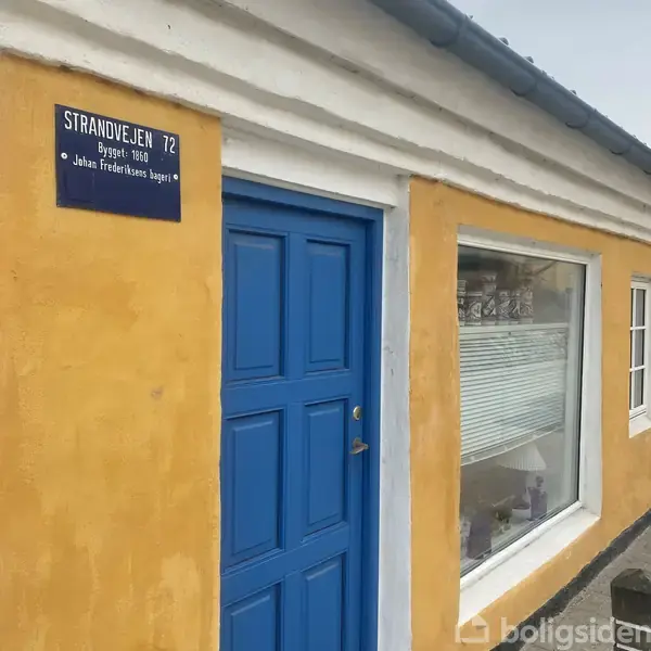 En gul murstensbygning med en blå dør og et stort vindue. Ovenfor døren er et skilt med teksten: "STRANDVEJEN 72 Bygget 1860 Johan Frederiksen's bageri".