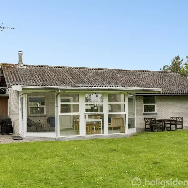 Et lille hus står stille i en velplejet have. Huset har store vinduer og en terrasse med bord og stole. Grønne træer omgiver området, der er fredfyldt og indbydende.