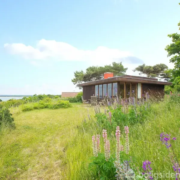 Et moderne sommerhus står roligt, omkranset af græs og vilde blomster. Det er placeret tæt på kysten med udsigt over havet, omkranset af frodig bevoksning.