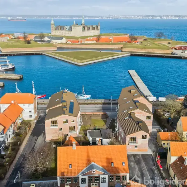 Kronborg Slot står majestætisk på en halvø omgivet af vand, mens farverige huse med røde tage ligger i forgrunden. Skibe ses nær kajen, og himlen er let overskyet.