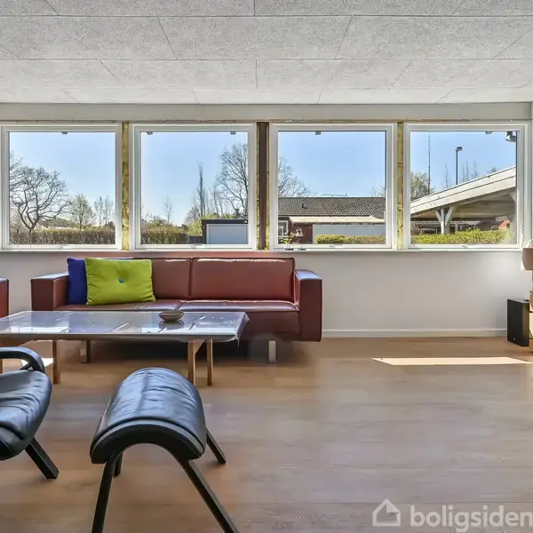 En stue med en brun lædersofa og farvede puder står ved et vindue. Et glasbord er placeret foran sofaen. Ved siden af står et TV og en moderne lænestol.
