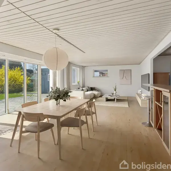 Spisebord med seks stole står i et lyst rum. Store vinduer viser en solrig have udenfor. En sofa og tv er i baggrunden, og der hænger en lampe over bordet.