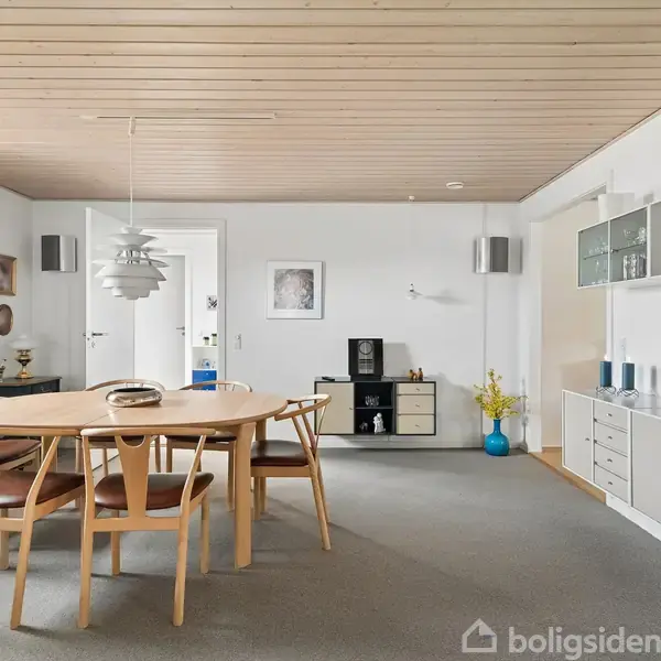 Køkkenområde med rundt bord og fire stole, prydet med en buket røde roser. Til venstre ses det åbne køkken, og i baggrunden er der adgang til en stue med kurvemøbler.