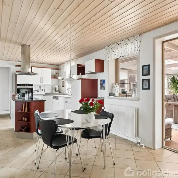 Sort spisebord med blomster og fire stole står i et lyst køkken med træloft og flisegulv. Køkkenet er åbent mod en stue med kurvemøbler og grønne planter.
