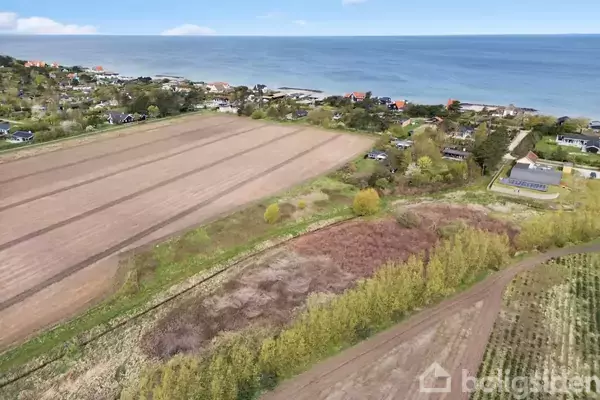 Mark med pløjede rækker ligger nær kysten. Omgivet af grønt buskads og et par huse. Havet strækker sig i baggrunden med blå himmel og lette skyer.