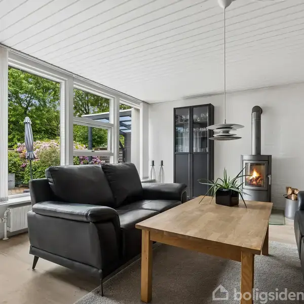 Sort lædersofa står ved et træbord i en stue, hvor en brændeovn opvarmer rummet. Store vinduer viser en terrasse med udsigt til en frodig have.