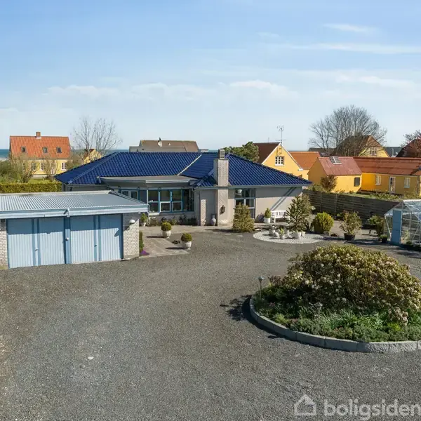 Et moderne hus med blå tagsten står stille omgivet af en stor grusbelagt gårdsplads. I baggrunden ses flere farverige huse og et drivhus i haven.