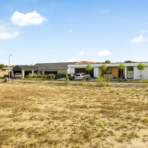 En moderne villa med fladt tag står stille ved siden af en indkørsel med biler, omgivet af en græsplæne og en lys sommerhimmel.