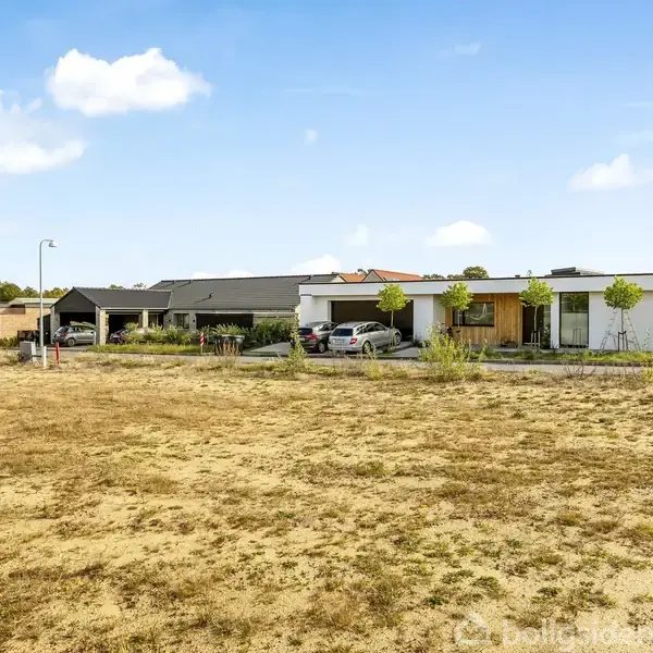 Flere moderne, flade tagehuse med store vinduer og græsplæner ligger i et beboelsesområde. I forgrunden ses en åben mark med sparsom vegetation. En klar blå himmel er i baggrunden.
