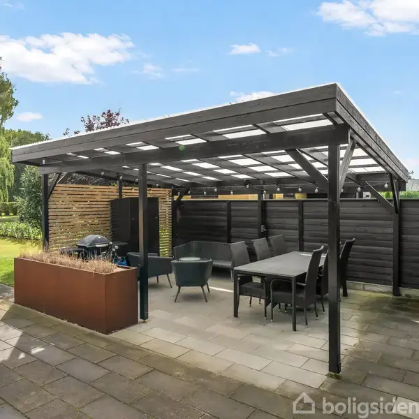 En overdækket terrasse med spisebord og stole står på en flisebelagt patio. I baggrunden ses en velplejet have med buske og træer under en blå himmel.