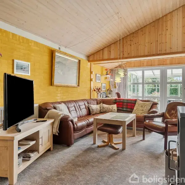 En stue med en brun lædersofa er hovedfokus. Sofaen står langs en gul væg, og på sofabordet er en plaid. Ved siden af står et TV på et træmøbel. I baggrunden ses store vinduer, som giver udsigt til en have. Væggene er dekoreret med malerier.