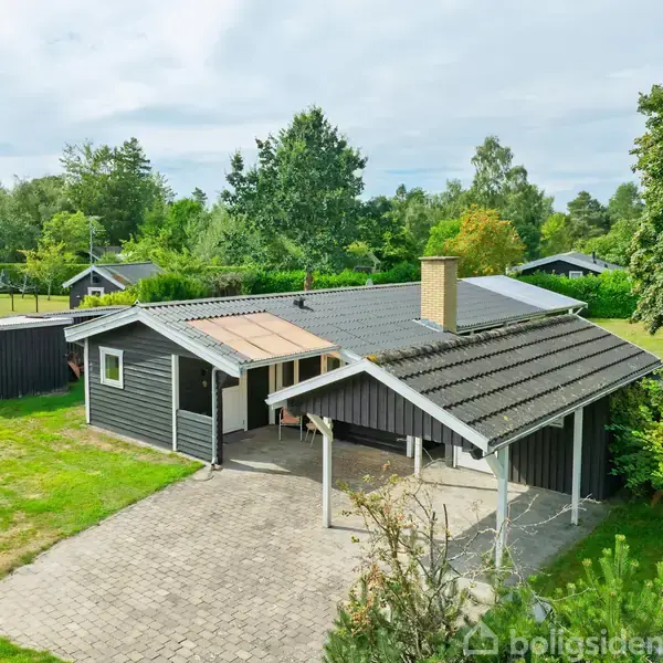 En sortmalet træhus står stille i en grøn have med buske og træer, omgivet af en flisebelagt gårdsplads. Huset har en skorsten og en carport.