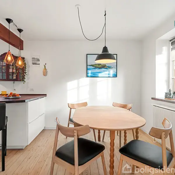 Rundt spisebord med fire stole står i et lyst køkken-alrum. En sort pendellampe hænger over bordet. På væggen hænger et farverigt billede, og der er store vinduer.