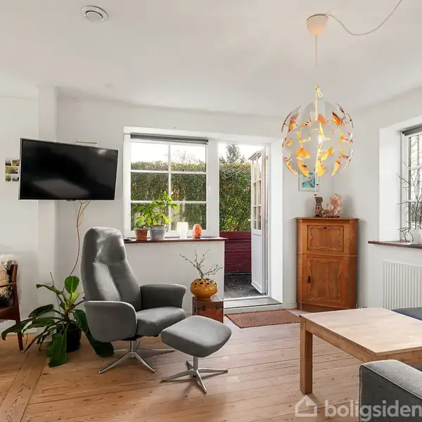 Stue med moderne lænestol og fodskammel foran fladskærms-TV. Rummet er lyst med store vinduer, træmøbler og grønne planter. En hængende lampe med orange dekorationer tilføjer farve.