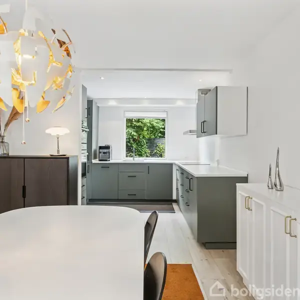 En moderne spisestue med rundt spisebord og dekorativ, hvid og gul lampe over. I baggrunden ses et lyst køkken med grå skabe og et vindue med udsigt til grønne træer.