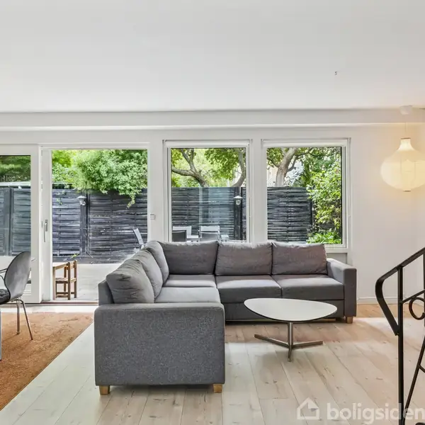 Gråt sofahjørne står i en stue med lyst gulv, foran store vinduer, der vender ud mod en træterrasse. Rundt bord ved siden af sofaen, spiseplads til venstre.