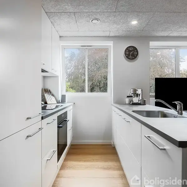 Køkken med hvide skabe og en sort bordplade. En indbygget ovn står til venstre. Der er en vask med armatur til højre og et vindue bagved. Der er ingen tekst.
