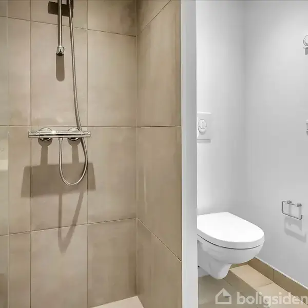 Bruser installeret på en væg af beige fliser i venstre side, med et tilstødende rum til højre, der har et toilet og toiletpapirholder på en hvid væg.