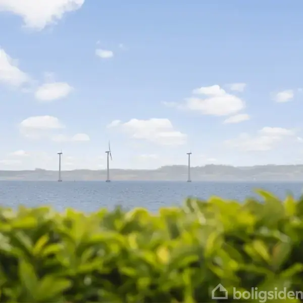 Vindmøller står stille over havet, indrammet af grønne buske foran. Blå himmel med spredte skyer i baggrunden skaber en rolig atmosfære.