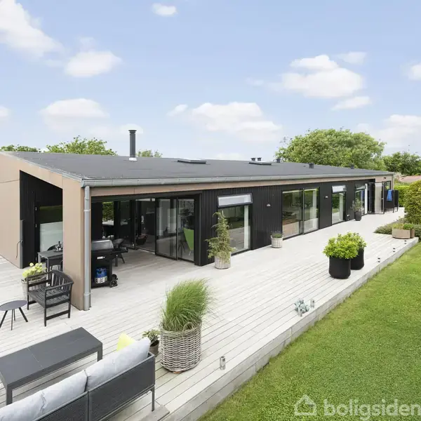En moderne bungalow står på en træterrasse omgivet af græsplæner. Terrasseområdet har havekrukker og udendørs møbler under en blå himmel med spredte skyer.