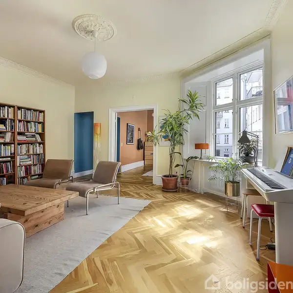 Stue med sofa, lænestole og træbord står på trægulv. Et fyldt bogreol og et keyboard er mod væggen. Store vinduer giver lys, omgivet af planter.