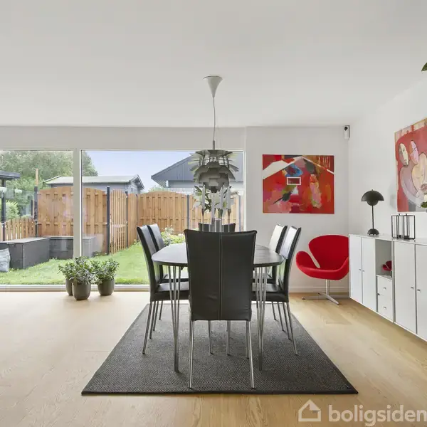 Et spisebord står på et tæppe med stole omkring. En stor glasdør åbner mod en have. På væggene hænger kunst, og en rød stol står ved siden af en hvid reol.