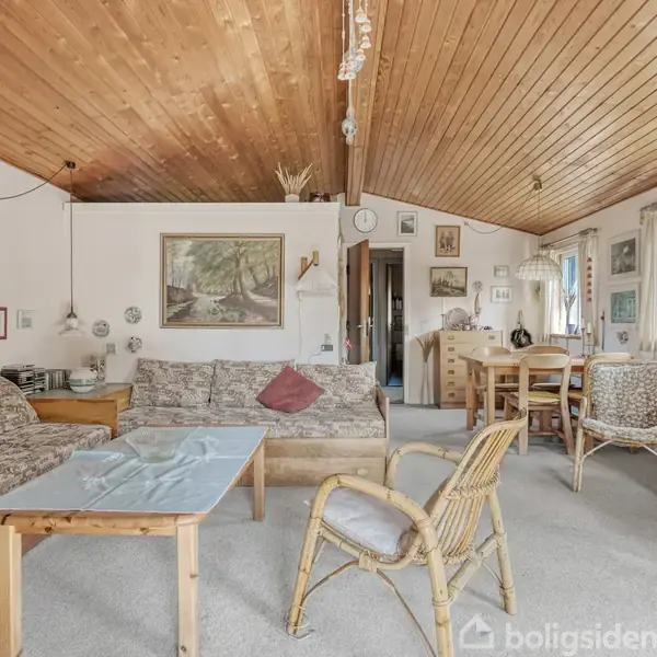 Rummet har en hyggelig stue med to sofaer og et sofabord, mange kunstværker på væggen, et træloft og store vinduer, der bringer lys ind.